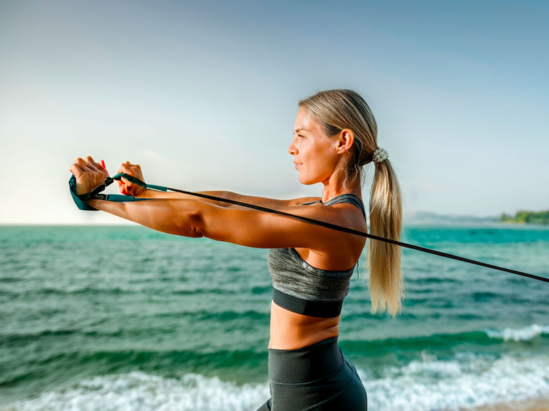 Žena cvičí s odporovou gumou u moře, procvičuje záda a trup.